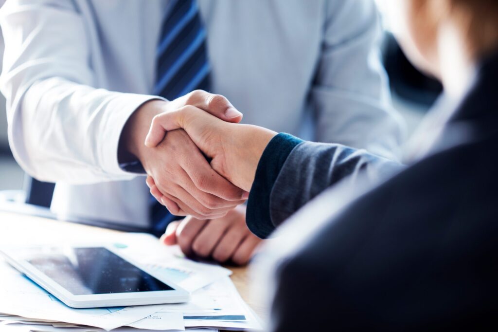 Two people in business attire shake hands over a desk with documents and a tablet, suggesting a professional agreement or meeting.