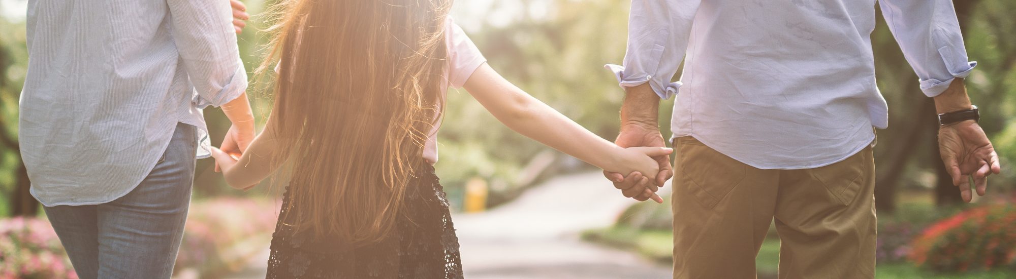 Three people walk hand-in-hand down a path, bathed in sunlight filtering through the trees. The central figure is a child with long hair, flanked by two adults. It's a serene moment reminiscent of the thoughtful family care seen in Brookfield Elder Law practices.