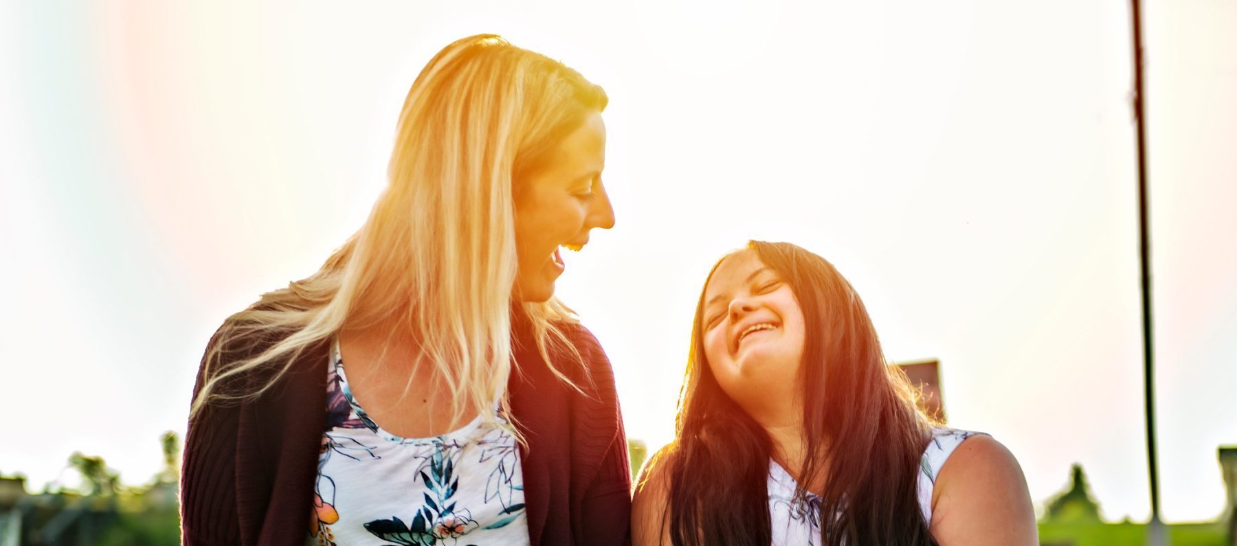 Two people, one blonde adult and one young girl with long dark hair, smile and hold hands outdoors in bright sunlight, both wearing floral-patterned tops.