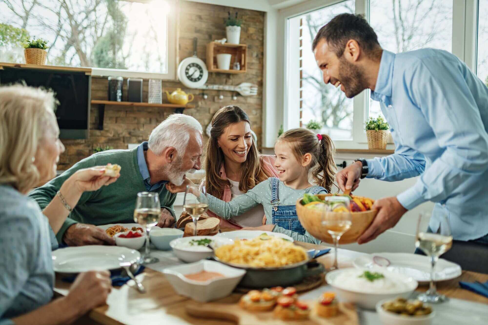 In a bright, cozy kitchen setting, a family enjoys a meal together at the dining table. A man serves salad while others smile and interact amid an array of dishes and wine glasses. It's moments like these that make up the cherished stories in life, akin to treasures safeguarded by Brookfield Elder Law.