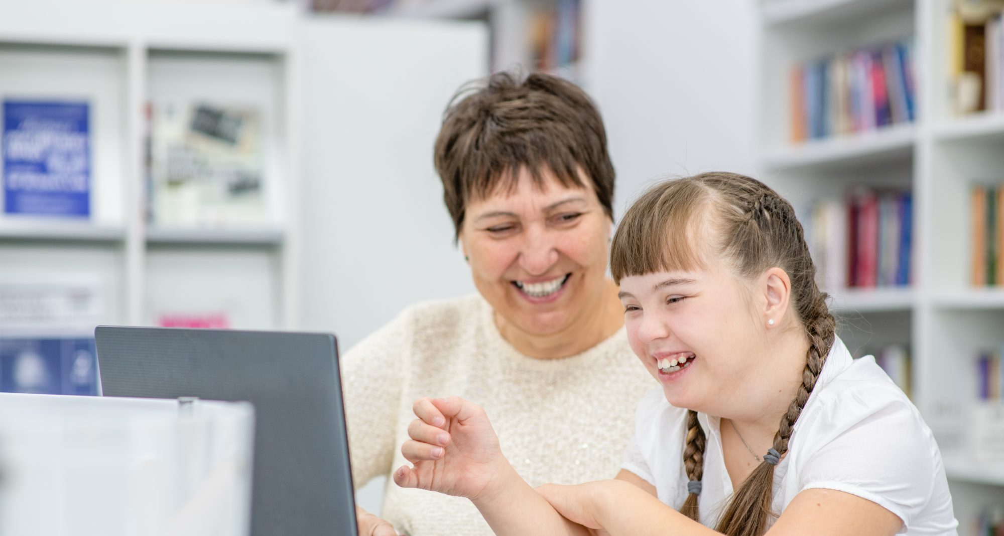 A smiling woman and a young girl with Down syndrome sit together at a table, looking at a laptop in a library or classroom setting.