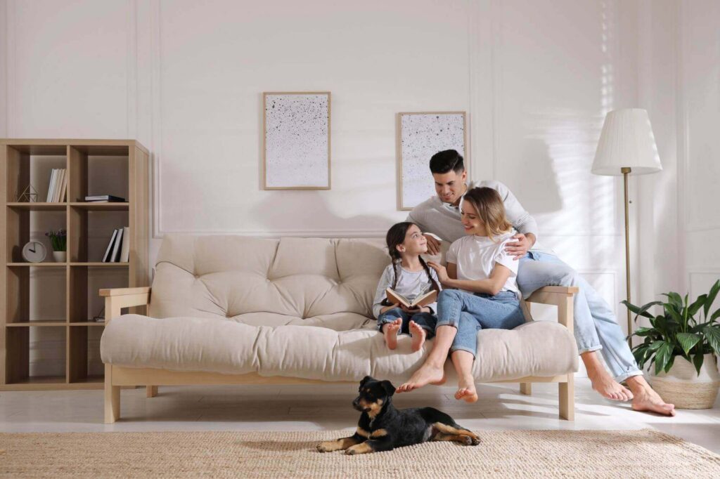 A family sits on a beige sofa in their well-appointed living room, with a young child immersed in a book. The dog lies contentedly on the floor, surrounded by homey touches like a bookshelf and potted plant. Framed artworks and plans from LaGrange Estate Planning complete the cozy scene.