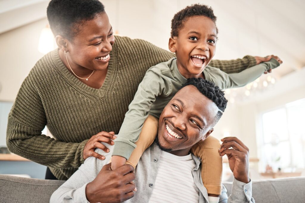 A smiling family, with a man carrying a child on his shoulders and a woman standing beside them, all indoors in their LaGrange Park home, discussing LaGrange Estate Planning.