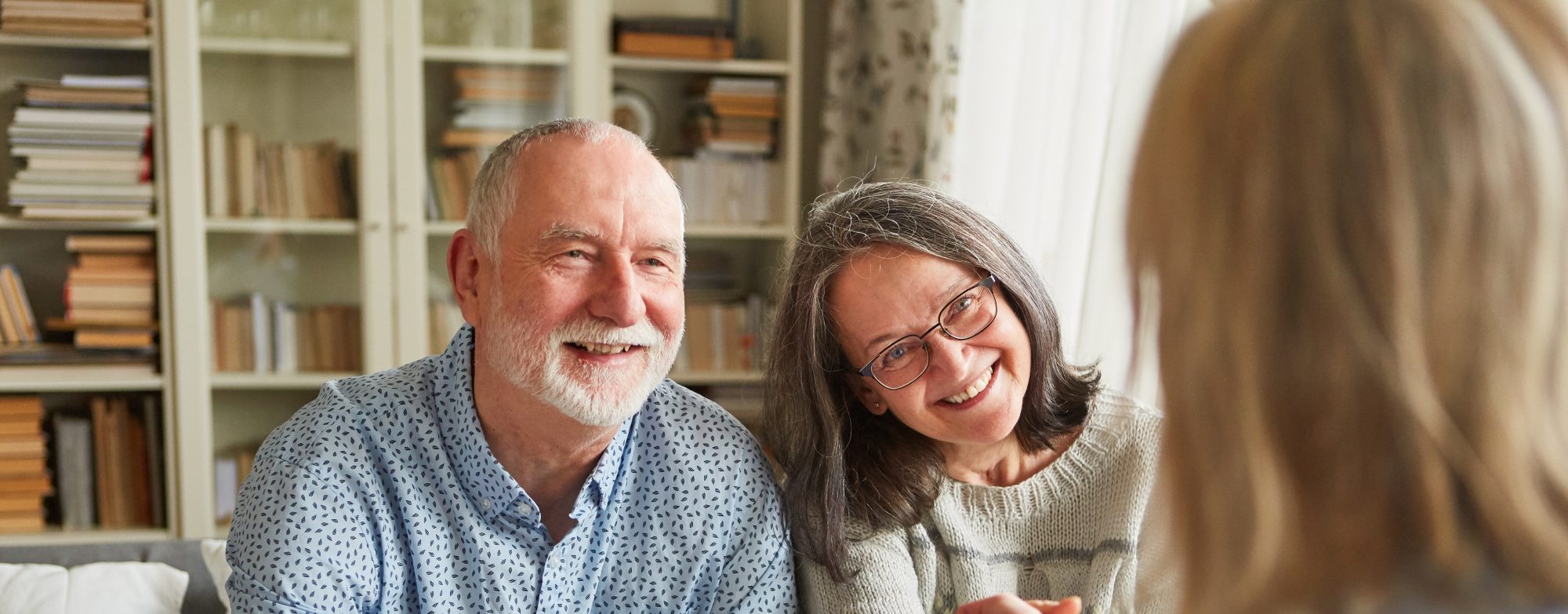An older couple sits on a couch, smiling, while the woman shakes hands with a person holding a clipboard in a home setting.