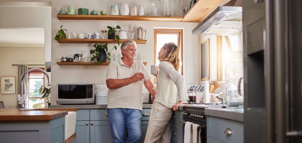 An older man and a woman stand in a modern kitchen with blue cabinets, talking and smiling while holding mugs, perhaps discussing advice from LaGrange elder law attorneys.