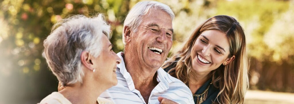 Three people sit outdoors, smiling and laughing together. An older man and woman sit next to a younger woman, enjoying a pleasant moment in the sun.