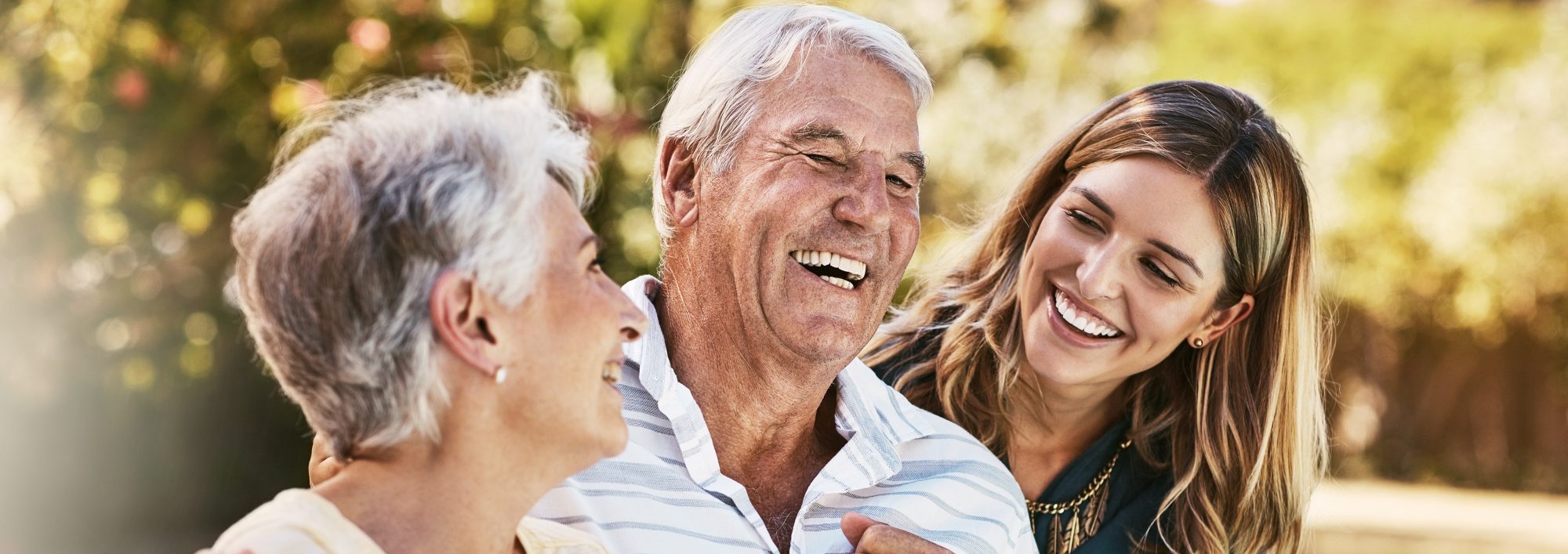 Three people sit outdoors, smiling and laughing together. An older man and woman sit next to a younger woman, enjoying a pleasant moment in the sun.