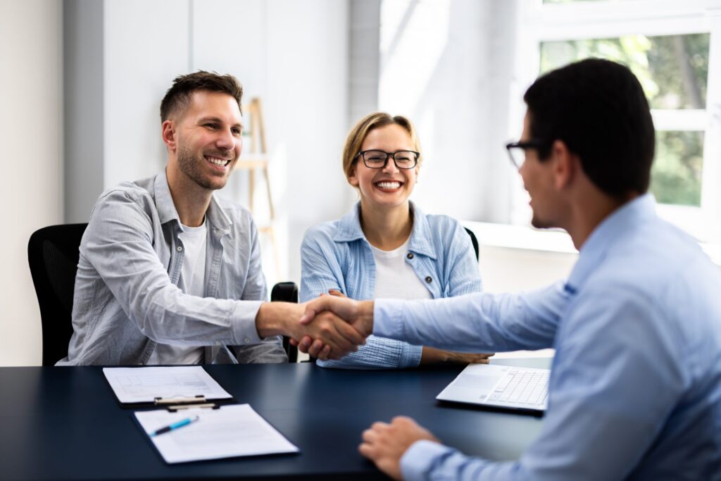 A man and woman smile as the man shakes hands with a Deerfield estate planning attorney across a desk in a bright office setting, with documents and a laptop on the table.