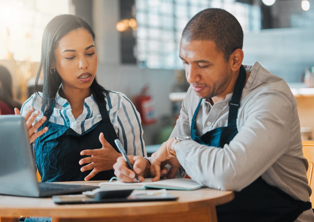 Two people wearing aprons sit at a table. One gestures while speaking, the other takes notes on Western Springs Estate Administration. A laptop is open in front of them, efficiently capturing their discussion.